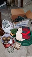 Photo of various Christmas decor items including a green plastic tree stand, a box and white bag of Christmas lights, disco ball ornaments, gold ball ornaments in a container, wood star-shaped piece, and fabric bags.