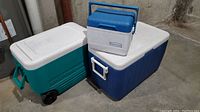 Photo showing two large rolling coolers, one turquoise Coleman and one dark blue Igloo, along with a smaller Rubbermaid lunch cooler on top of one cooler.