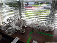 Photo showing the full lot of crystal items including bowls, vase, pitcher, and jars arranged in natural light on a table near window blinds, clearly showing detailed cut and etched patterns.