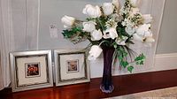 Photo of two floral-themed art prints in square silver frames alongside a purple-tinted art glass vase with white faux flowers bouquet.