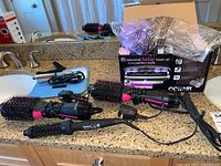 Photo showing three hair styling tools including black and pink Revlon curling brush, curling iron, and a Conair curling roller set with original box in background.