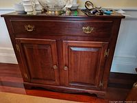 Front angle of dark wood Harden sideboard table with brass handles and paneled cupboard doors. Various household items on top not included.