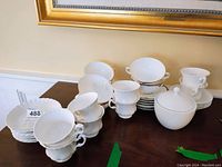 Image showing grouped white porcelain teacups and saucers with lidded sugar bowl on wooden table under a framed painting.