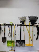 Garage wall mounted tool rack with various shovels, rakes, and mops hung and marked with green tape
