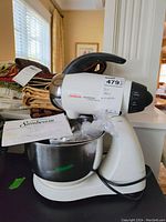 Vintage cream and black Sunbeam MixMaster stand mixer on black surface with stainless steel mixing bowl attached. Instruction manual and plastic-wrapped attachments placed inside the bowl. Background shows folded fabrics on table near window.