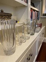 Photo of nine clear glass vases of varying shapes and sizes arranged on a white cabinet shelf with books in the background.