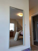 Full view of the rectangular framed mirror hung on a wall showing reflection of a bedroom interior including furniture and lighting fixture.