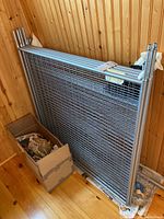 Side view of folded metal drying rack leaning against a wood-paneled wall with box of parts on floor