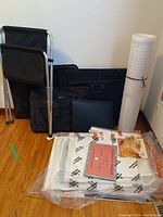 Photo of assorted art carrying portfolio bags, rolled papers, and folded chrome art holder chair leaning against the wall on wood floor.