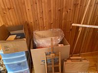 Five wooden easels displayed along a wood-paneled wall with cardboard boxes containing papers and mats, and a blue plastic storage tote below.