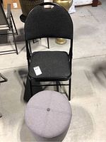 Front view of black metal folding chair with padded dark grey fabric seat and backrest. Round grey fabric storage ottoman in front.