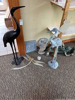 Full view of lot showing large black metal crane, lighter metal crane garden stake, stepping stones, owl figurines, dragonfly stake, ladybug figurine, and fairy figurine on shelf.