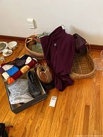 Photo showing lots of items grouped on floor including knit Afghan, hiking boots in box, tulip bag, two woven baskets, purple coat and button-up shirt.