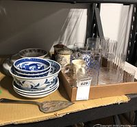 Shelf with stacked Noritake Ambiance plates (2 sizes), Asian-themed blue and white bowls, various clear glasses with vertical stripes, cream-colored mugs, a small decorative jar with lid, and a metal cake server.