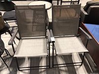 Pair of folding patio chairs with brown armrests, light beige mesh fabric seats and backs, black metal frames.