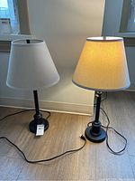Pair of black metal table lamps with beige fabric shades placed on wood flooring near window. Both lamps have cords with plugs visible.