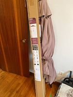 The folded patio umbrella leaning against the wall alongside the packaging box with product info visible. Shows umbrella fabric tied closed and cardboard packaging labeled 'Aberdeen Round Market Umbrella.'