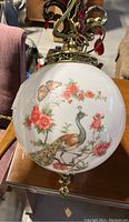 Close-up photo of white glass globe showcasing detailed peacock and butterfly painted motif with red flowers and green leaves.