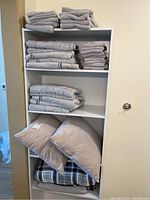 Shelf with stacked gray and beige towels, two beige throw pillows, and folded blankets including a plaid one