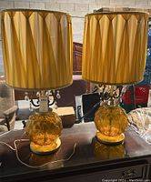 Photo showing both vintage lamps with yellow-orange glass bases, pleated fabric shades, metal details, and hanging prisms, placed on a table in an indoor warehouse setting.