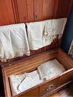 Nine antique linens displayed in and on a carved wooden chest, showing lace trims, embroidery, and aged white linen fabric.