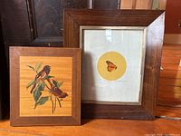 Two framed artworks shown side by side on wooden floor: butterfly print on right, bird wood inlay on left