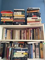 Books stacked on two shelves and a pile on top of a shelving unit, mostly soft covers with visible titles