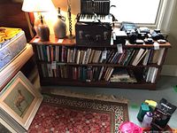 Photo showing pine bookshelf with two shelves filled with various books, placed against a wall under a window. Other objects such as lamps and miscellaneous items are on and around the bookshelf but not included in the lot.