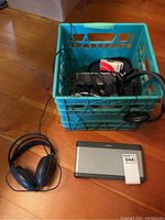 Blue storage crate with tangled headphone wires and accessories inside. Outside the crate, the Bose SoundLink Mini Bluetooth speaker and a pair of headphones with black headband and wired connection are placed on wooden floor.