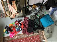 Photo showing assorted camping and outdoor gear including bags, jackets, sleeping bag, and various small items arranged on a carpet and floor.