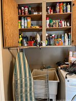 Photo of wall cabinets filled with various cleaning supplies above laundry sink and washing machine. Iron and batteries visible hanging and placed nearby. Ironing board leaning against wall, metal drying rack in foreground.
