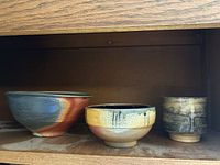 Three pottery bowls arranged side-by-side on wooden shelf showing size differences and glaze styles.