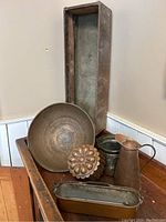 Six copper pieces arranged on wooden surface: tall tray, bowl, jelly mould, pot, jug, and long narrow tray.