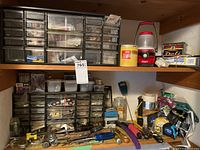 Wide view of two sets of black plastic storage drawers filled with small hardware and hand tools placed on a wooden shelf with additional hardware on shelves above.