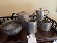 Five-piece hammered pewter set arranged on a wooden surface including teapot, coffee pot, creamer, sugar bowl, and a large bowl. All have textured, hammered finishes.