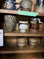 Photo showing shelves with various pottery pieces including mugs with handles and cups without handles with varied glazes and patterns.