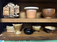 Photo showing two shelves with six pottery items; front houses the decorative house-shaped vase and various small bowls in different colors and finishes.