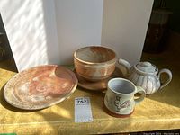 Photo showing the full set of pottery items: plate, planter with saucer, cup, and teapot on a yellow surface with sunlight.