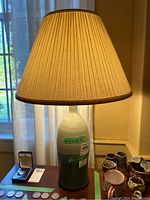 Front view of pottery table lamp with shade on a wooden surface near window. Lamp is lit and shade is beige with brown trim.