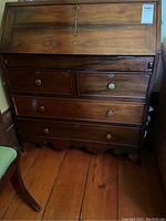 Front view of wooden secretary desk with drop front closed, showing four drawers with round brass knobs and keyhole escutcheons, on wooden floor.
