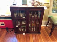 Front view of small wooden display cabinet showing glass pane doors with vertical wooden pickets, placed on hardwood floor.