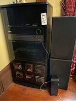 Photo showing the black entertainment cabinet with glass front containing stereo components including a Harman Kardon receiver, Pioneer compact disc player, and multiple CDs stored in bottom compartments. Floor speakers are positioned beside the cabinet.