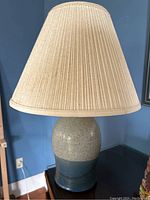Full view of the pottery table lamp with pleated beige shade on a wooden surface against a blue wall.