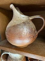 Side view of round pottery jug showing handle and spout with brown and beige glazing.