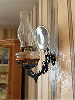 Front view of the oil lantern wall sconce showing the glass chimney, oil reservoir with reddish tint, and cast iron mount with reflector.