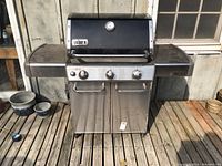 Front view of stainless steel Weber BBQ with side tables and closed cabinet doors, showing the propane tank inside and control knobs on front panel