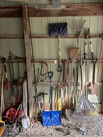 Photo of assorted yard tools hanging on a wooden rack including shovels, rakes, broom, and blue plastic snow shovel.