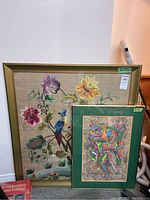 Photo showing two framed artworks; the larger textile art depicting birds and flowers with a gold frame, and a smaller green-framed vibrant painting with birds and colorful patterns.