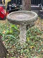 Full view of one cement birdbath showing basin and decorative sculpted pedestal base in fallen leaves and outdoor setting.