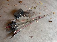 Full view of multiple fishing rods with attached reels laid on floor next to black metal tackle box and fishing net with telescoping handle